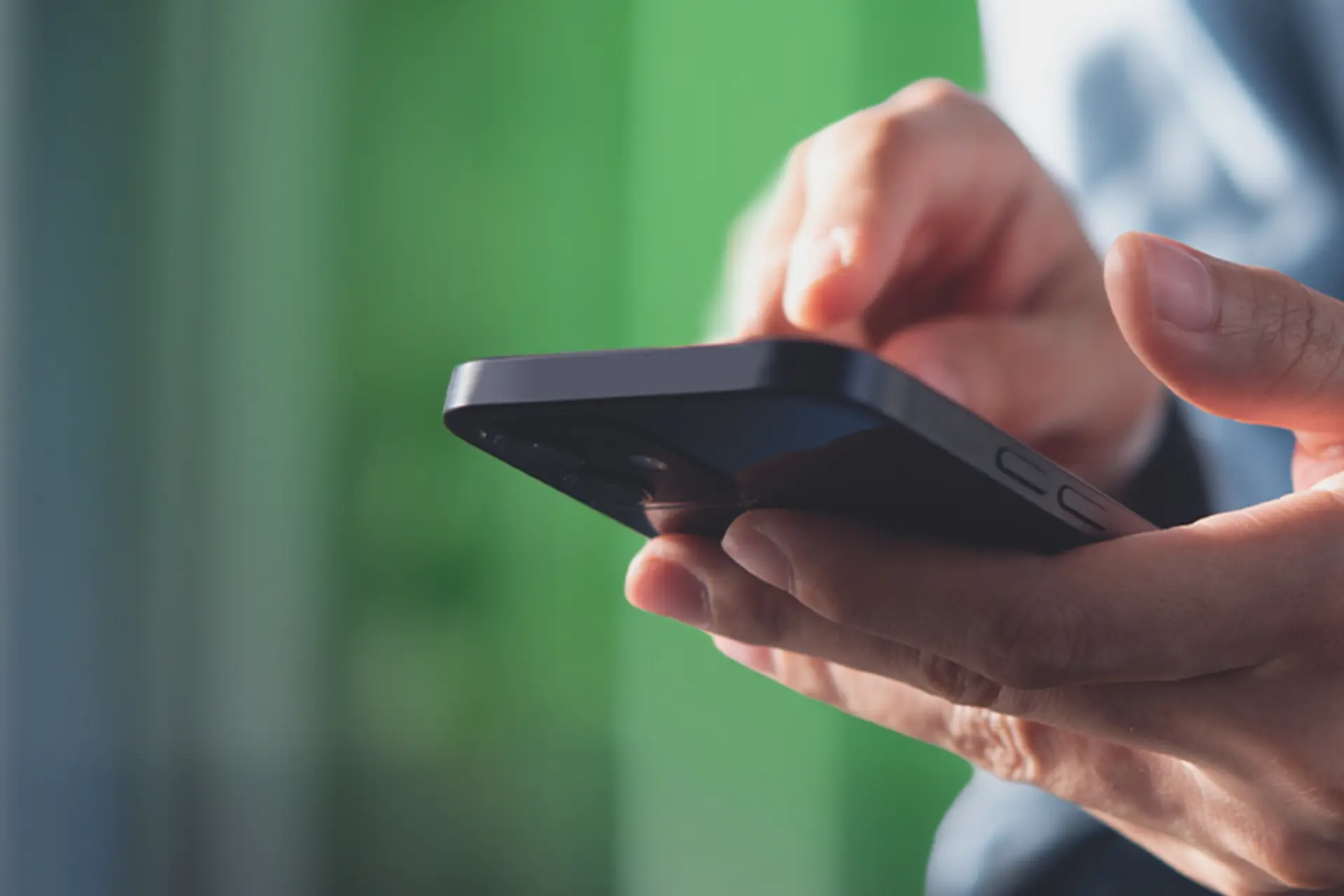 Primer plano de manos sosteniendo un smartphone negro mientras interactúan con la pantalla; fondo luminoso desenfocado en verdes y azules.