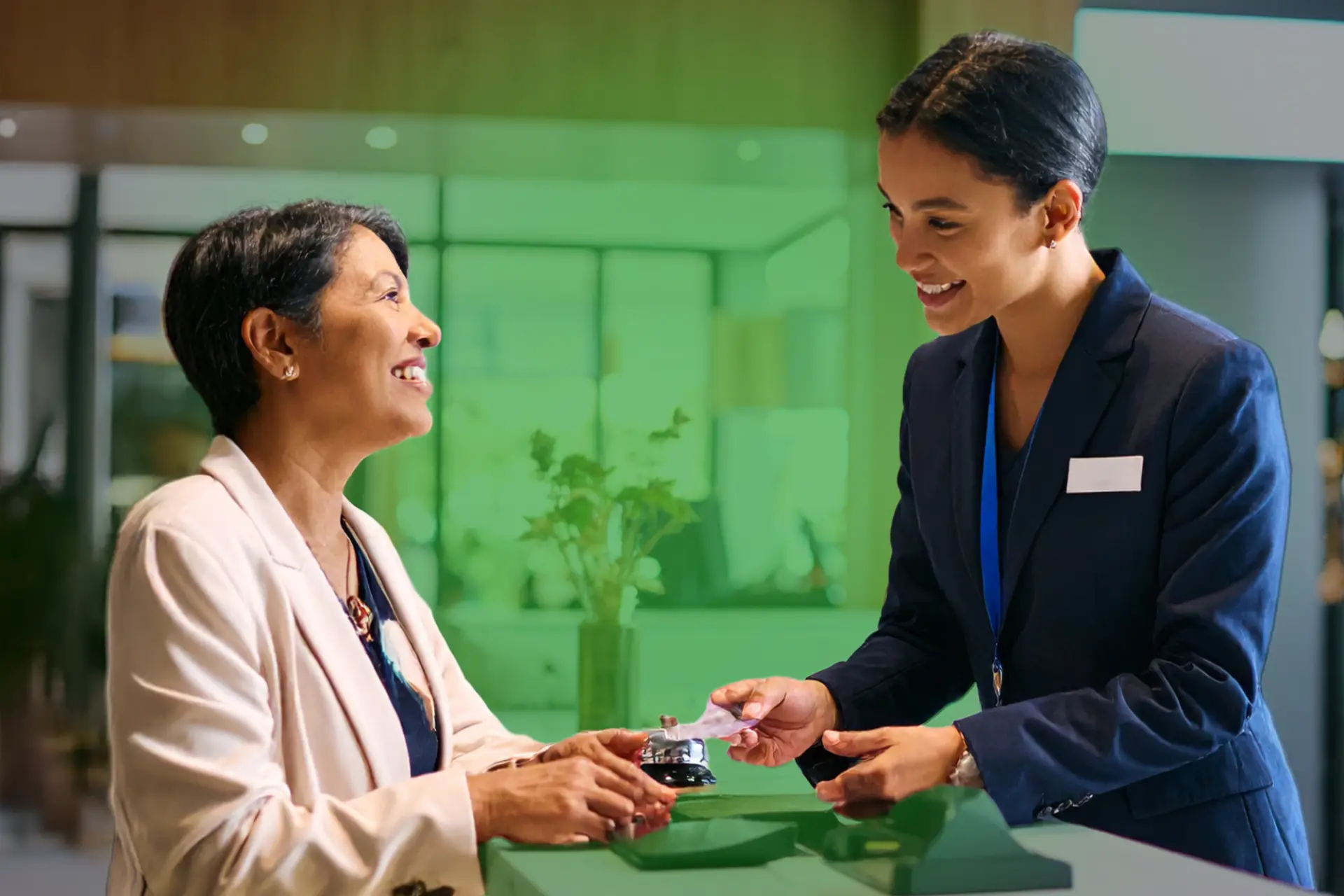 Recepción de hotel con dos mujeres sonriendo: recepcionista en traje azul entrega una tarjeta o recibo a visitante sentada con blazer claro; mostrador moderno con campana y ambiente interior bien iluminado.