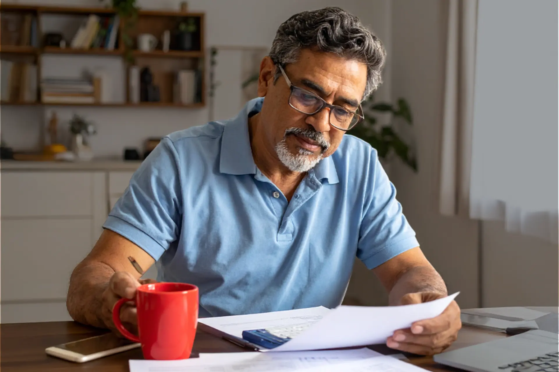 Hombre de mediana edad con gafas leyendo documentos en una mesa de madera, con taza roja, smartphone y calculadora; ambiente doméstico bien iluminado.